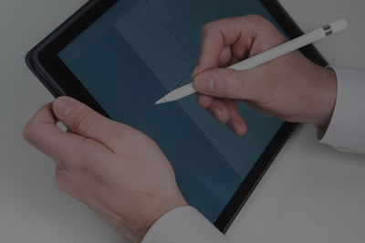 A mão direita de uma pessoa branca segura um tablet e a mão esquerda usa caneta especial para escrever na tela.